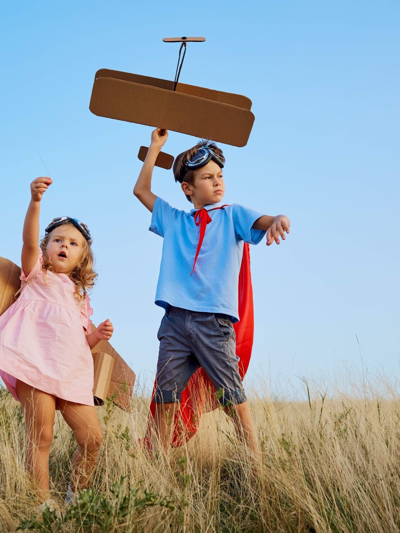Two preschool kids wearing costumes, playing with a cardboard airplane outdoors
