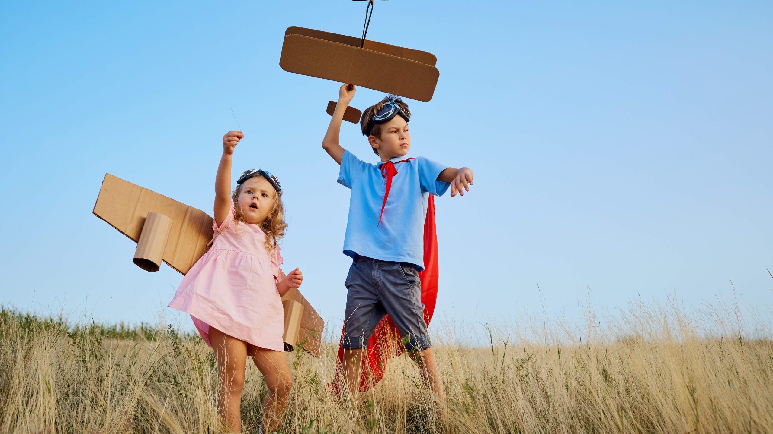 Two preschool kids wearing costumes, playing with a cardboard airplane outdoors